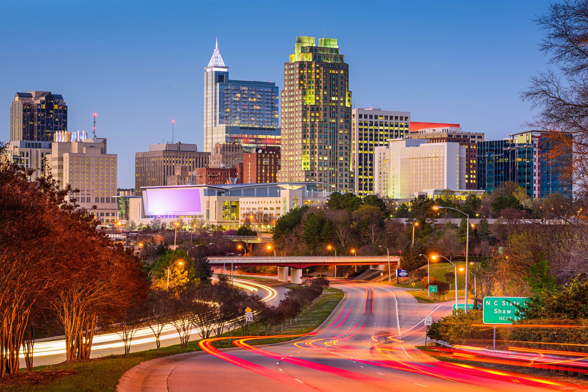 Downtown Raleigh skyline at dusk, with traffic light trails on the highway below